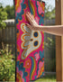 A rectangular polyester doormat with a vibrant, psychedelic design on a hot pink background. In the center is a cute, cartoonish ghost in off-white with yellow and orange outlines; it has large, dark, expressive eyes. The ghost is surrounded by a maximalist pattern of 70s-style floral motifs and puffy clouds in shades of teal, blue, orange, and light pink. Two small, colorful rainbows arc over the ghost's shoulders. The doormat has a durable, textured surface and is framed by a sleek black trim.